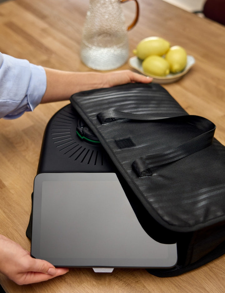 Person opening a black suitcase on a wooden table with a plate of fruit in the background.